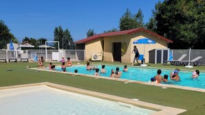 Una piscina con gente nadando, un puesto de salvavidas y un pequeño edificio de techo rojo.