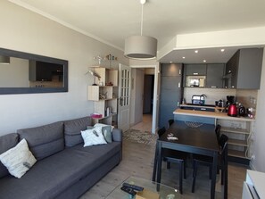 Ein modernes Wohnzimmer mit einem grauen Sofa, einem Esstisch mit Stühlen und einer Küchenzeile inklusive Kaffeemaschine.