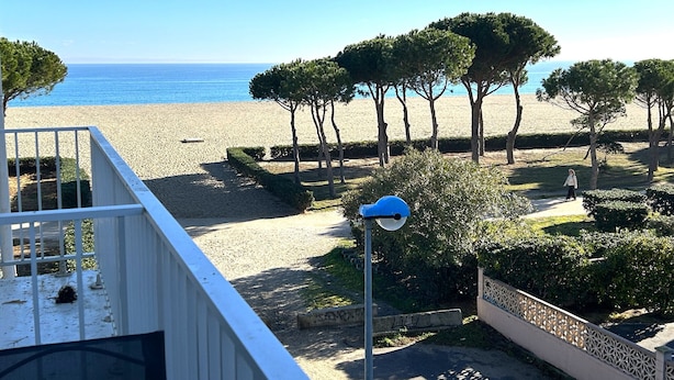 Vista a la playa desde un balcón, con cielo despejado, arena y una hilera de árboles.