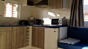 Una kitchenette compacta con microondas, cafetera y tostadora. Hay un refrigerador pequeño y armarios de madera. Un sofá azul está ubicado cerca de la ventana, con persianas parcialmente bajadas.
