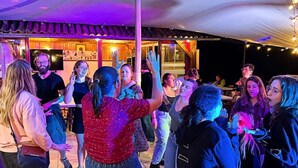 Eine Gruppe von Menschen tanzt auf einem Freigelände mit überdachter Sitzgelegenheit und einer Bühne im Hintergrund.
