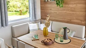 Ein Essbereich mit einem Holztisch, beigen Stühlen und einer Bank mit Kissen. Zwei Fenster mit Vorhängen, eine Vase mit Trockenpflanzen und ein Krug auf dem Tisch sind ebenfalls vorhanden.