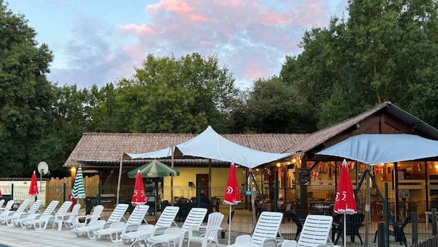 Ein Poolbereich mit Liegestühlen und Sonnenschirmen, ein Gebäude mit Holzbalkonen und einem Ziegeldach, sowie eine natürliche Kulisse aus Bäumen.