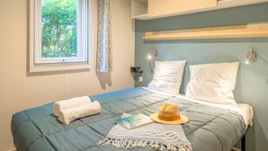 A bedroom with a bed, a window with a view of greenery, a bedside table, and a wall-mounted shelf. - Mobile home with terrace, 3 bedrooms, for 6 people (Rivedoux-Plage)
