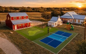 A red barn, a white house, and a tennis court with a green field and trees in the background. - 6K SqFt Hill Country Estate- Creek, Barn, Sport Court (Fredericksburg)