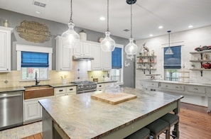 A kitchen with white cabinets, a granite countertop, a central island, hanging pendant lights, and a window with blue blinds. - 6K SqFt Hill Country Estate- Creek, Barn, Sport Court (Fredericksburg)
