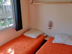 A room with two beds, each with an orange bedspread and white pillows. There is a window with a curtain, and a small shelf above the beds. - 3-bedroom mobile home with terrace in Collias (Collias)