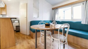 A compact, modern room with a kitchenette, a dining area with a round table and two chairs, and a teal sofa against a window with curtains. - Mobile home 4 pers. with terrace & grill (Rivedoux-Plage)