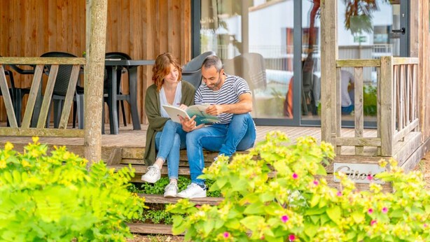 Two people sitting on wooden steps outside a building with a glass door, surrounded by greenery and a wooden fence. - Mobile home 4 pers. with terrace & grill (Rivedoux-Plage)