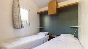 A small, simple room with two single beds, a window with curtains, and a wooden shelf above the beds. - Mobile home 4 pers. with terrace & grill (Rivedoux-Plage)