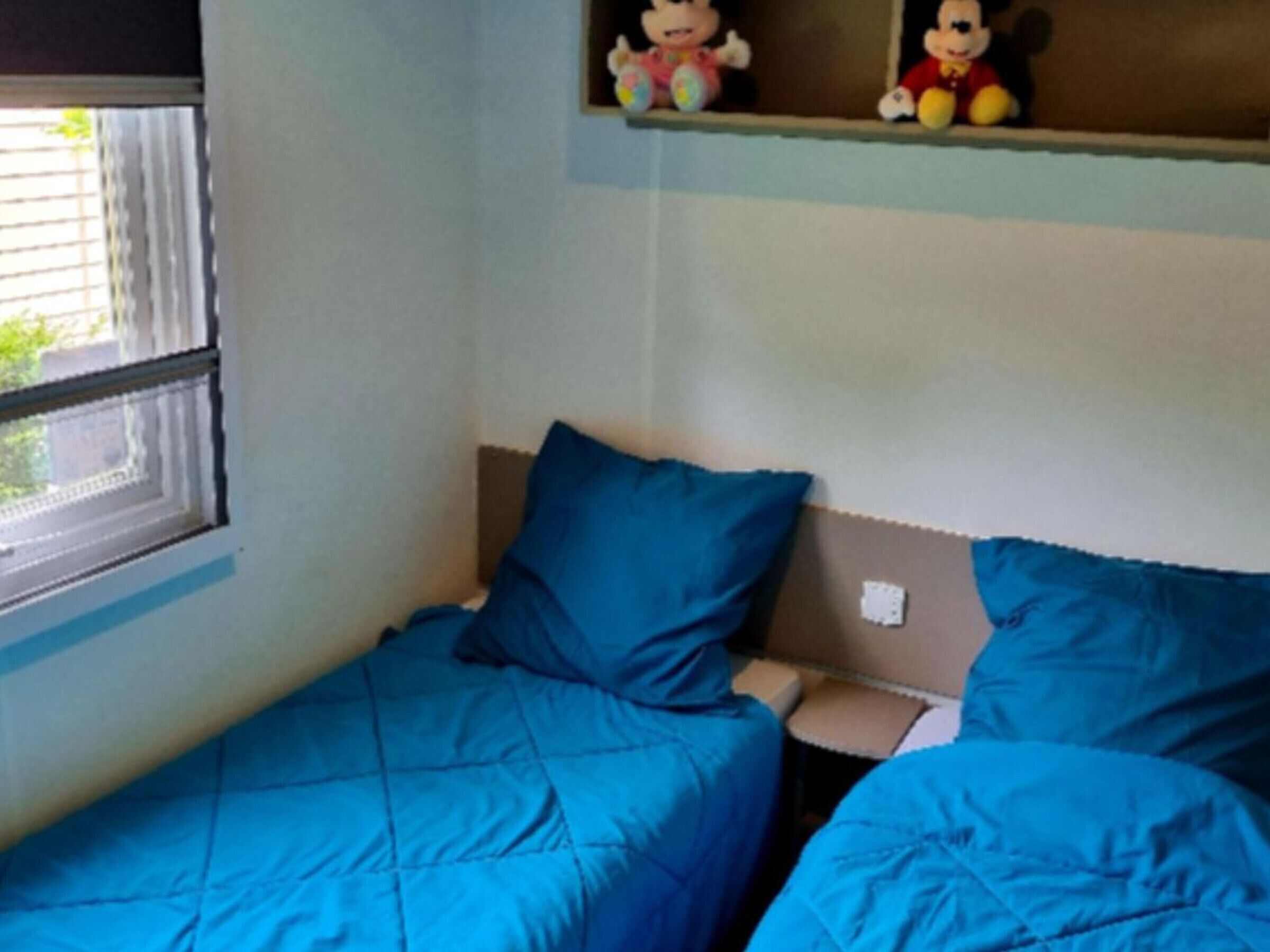 A small, single-bed room with a blue bedspread, a window with blinds, and a shelf with two Mickey Mouse figurines.