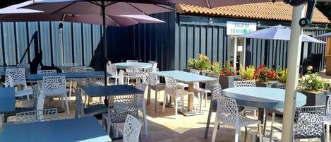 An outdoor seating area with tables and chairs under umbrellas, a sign reading "SALLE DE SEMINARE," and a building with a tiled roof in the background.