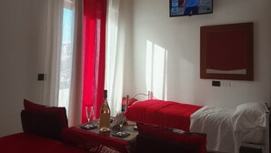 Et hotelværelse med to senge, rødt sengetøj, et fjernsyn på væggen og et bord med en flaske og glas.