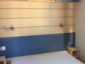 Ein Zimmer mit einem Bett, einem kleinen hölzernen Hocker und einer Wand mit horizontalen Linien und einem blauen Streifen.
