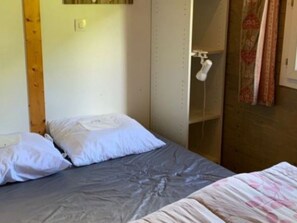 Una cama individual con colchón gris y almohadas blancas, cabecera de madera y pared blanca con toma de corriente.