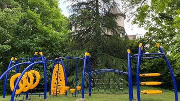 Une aire de jeux avec des équipements bleus et jaunes, entourée d’arbres et avec un bâtiment visible au loin.