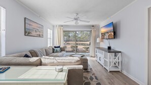 Una sala de estar moderna con un sofá seccional, una mesa de centro de vidrio, un televisor montado en la pared y vistas al exterior a través de ventanas amplias.