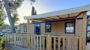 En träkonstruktion med ett däck och ett taköverbyggt område, troligen en stuga eller ett litet hus, omgiven av träd och andra liknande byggnader i en camping- eller husvagnsområde.