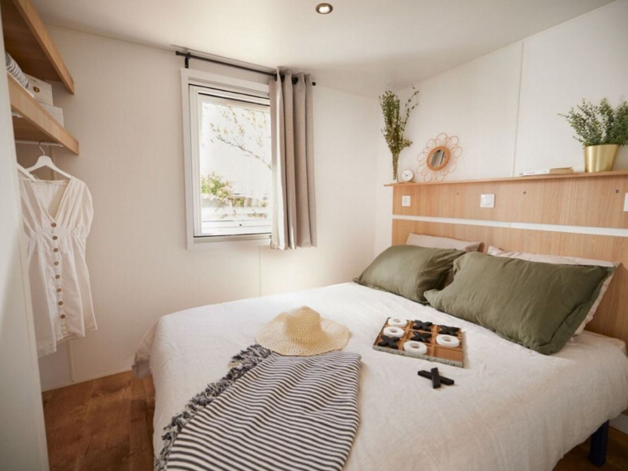 A bedroom with a bed, a window, a shelf with plants, and a hanging garment.