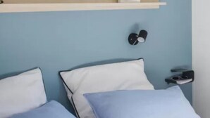 A bedroom with a bed, pillows, a nightstand, a wall-mounted lamp, and a shelf above the bed. - Mobile home for 2 with terrace (Sandrans)