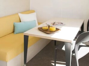 Een kleine, lichte kamer met een geel bankje, een witte tafel en een grijze stoel.