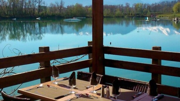 Eine Holzterrasse mit Tischen und Stühlen mit Blick auf einen stillen See und Berge im Hintergrund.