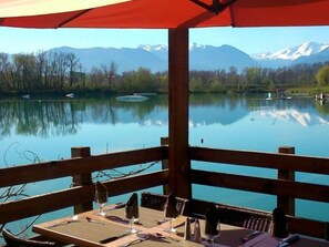 Une terrasse en bois, agrémentée de tables et de chaises, offre une vue sur un lac paisible, avec des montagnes en toile de fond.