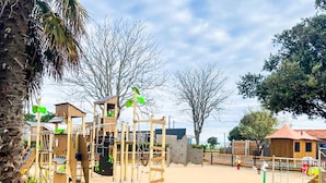 Un terrain de jeux doté de diverses structures ludiques, entouré d’une clôture en bois et de palmiers.