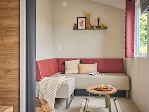 A cozy corner with a red sofa, a small round table with a tray of pastries, and a wicker basket. - Mobile Home 6 pers with Terrace - New Style! (Concourson-sur-Layon)