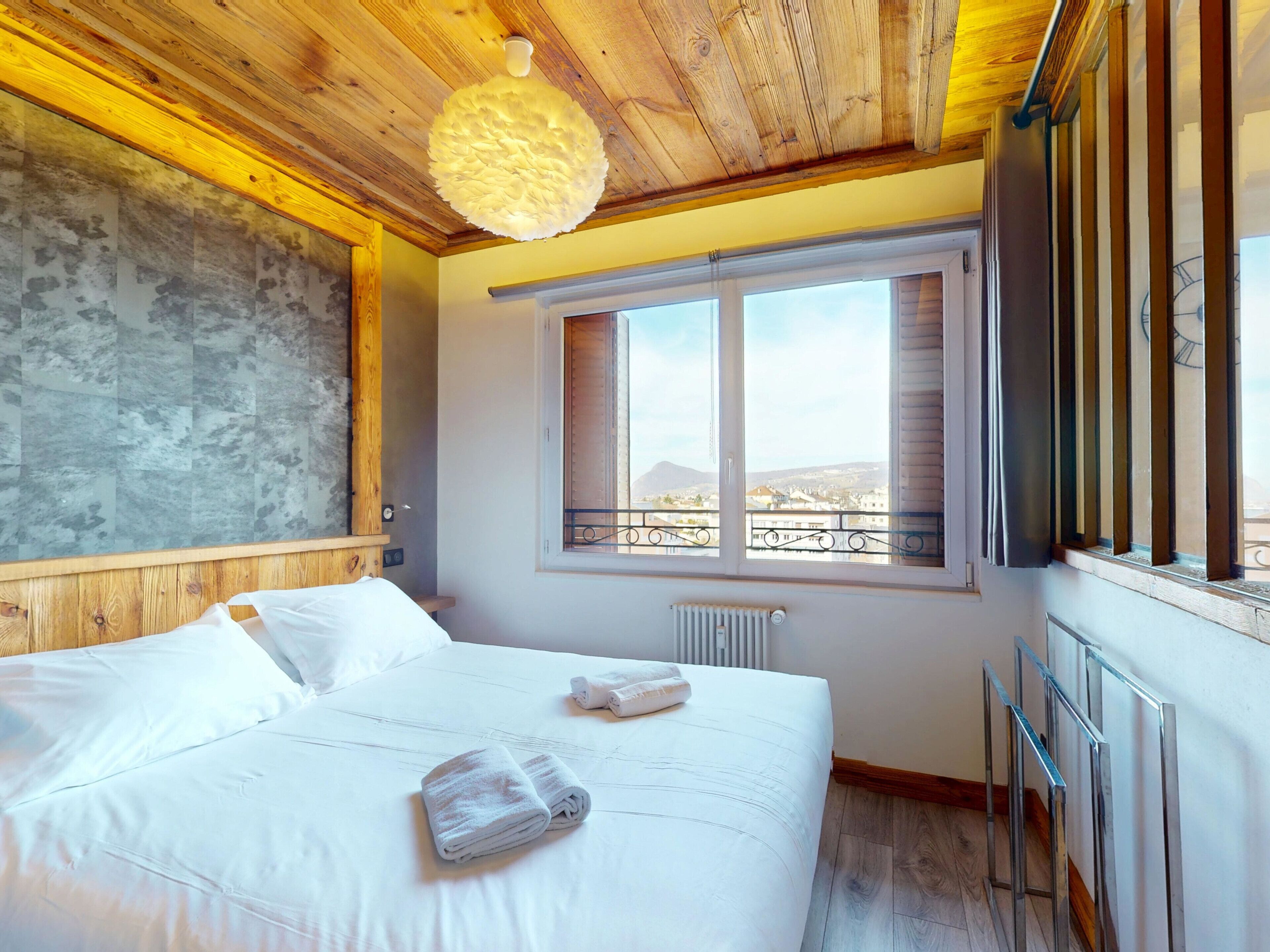 Ein Schlafzimmer mit Holzboden, einem großen Fenster mit Blick und einem Bett mit weißer Bettwäsche, sowie einem Heizkörper.