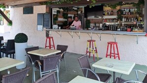 An outdoor café with wicker tables and red stools, a bar area with shelves of bottles, and a person working behind the bar. - Mobile home for 6 with terrace (Laurens)