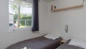 A room with two beds, a window with curtains, a shelf with books, and a car visible outside. - Cozy mobile home for 4 people with fully-equipped kitchen (Les Aires)