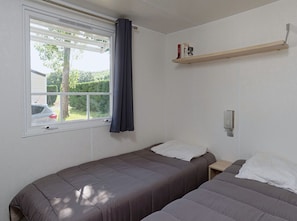 A room with two beds, a window with curtains, a shelf with books, and a car visible outside. - Cozy mobile home for 4 people with fully-equipped kitchen (Les Aires)