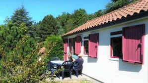 A white building with a red-tiled roof and pink shutters, surrounded by greenery.