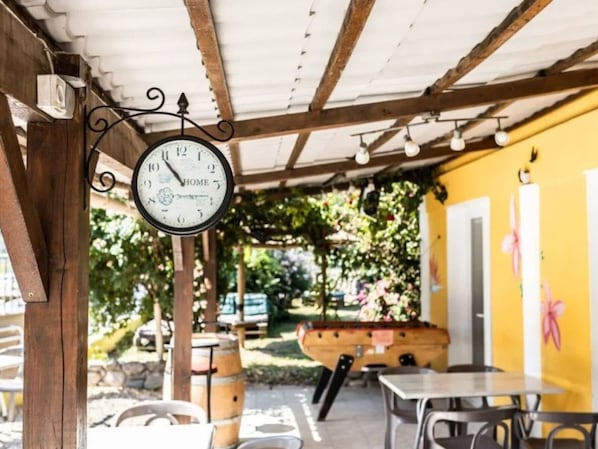 Un espace extérieur abrité avec des places assises, agrémenté d’une horloge suspendue, de poutres en bois et d’un mur jaune.