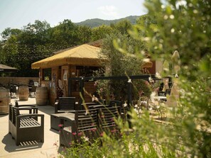 An outdoor seating area with tables and chairs, a thatched-roof structure, and a view of a forested hill. - Mobile Home 6 Pers, Terrace (Savignac-les-Ormeaux)