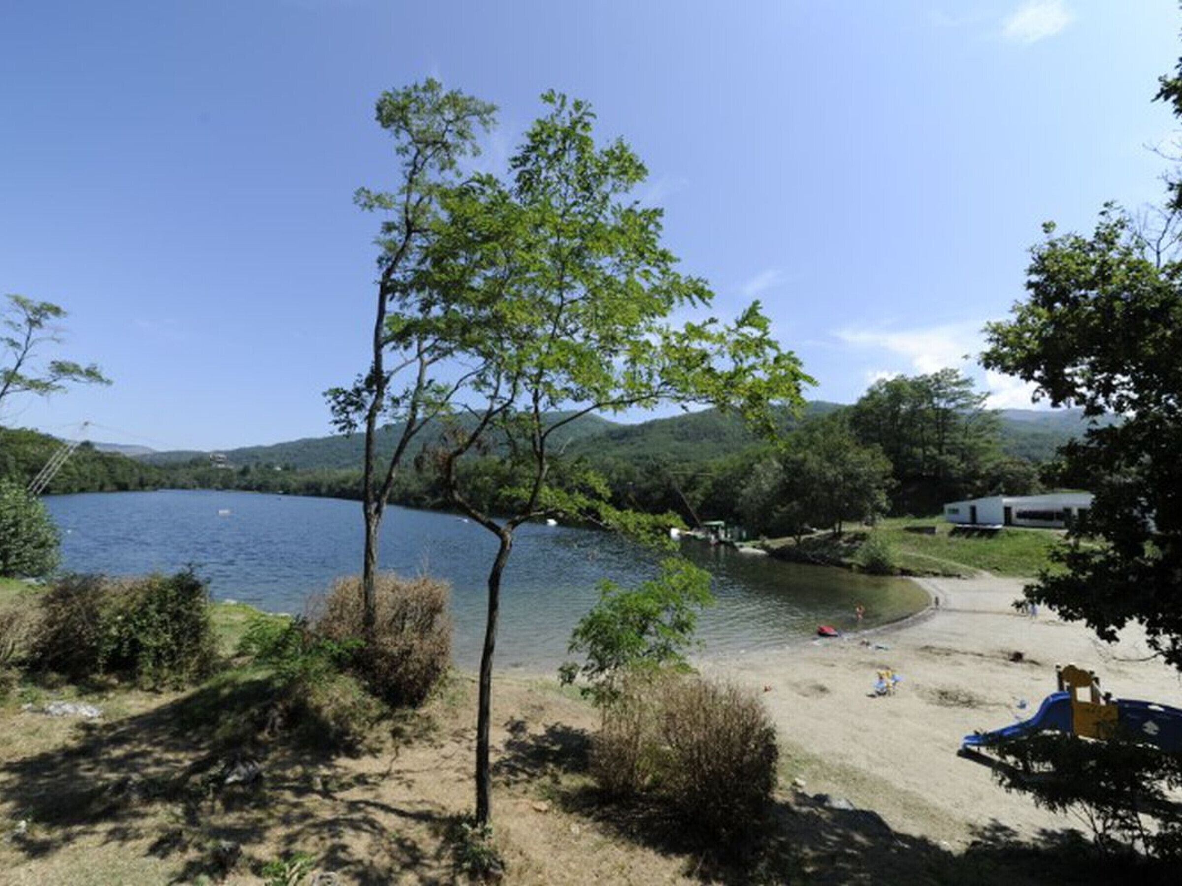 Un camping frente a un lago, con playa de arena, un muelle pequeño y una caravana.