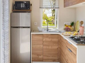 Une cuisine compacte équipée d’un four à micro-ondes encastré, d’un réfrigérateur en acier inoxydable, de meubles de rangement en bois, d’une fenêtre donnant sur un espace verdoyant et d’une cuisinière à gaz.