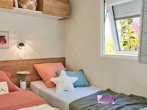 Une chambre avec deux lits, une fenêtre donnant sur des arbres, une étagère murale agrémentée d’objets décoratifs et un applique murale.