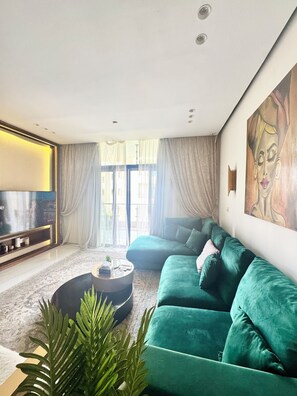 A modern living room with a green sofa, a flat-screen TV, and a large window with curtains. - Beautiful Modern 2-bedroom apartment with AC in tranquil New Cairo (New Cairo)