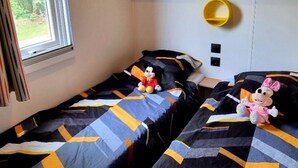 Zwei Einzelbetten mit gemusterten Bettwäsche, ein Fenster mit Vorhängen, ein Ablagefach mit Handtüchern und ein kleiner gelber Korb.