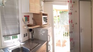 Une cuisine compacte équipée d’un évier, d’une cuisinière et d’un four à micro-ondes. On y trouve une fenêtre avec des stores, une porte donnant sur un balcon et une petite étagère avec une machine à café.