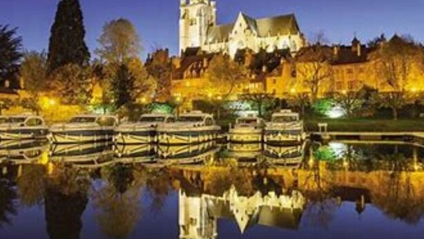 Ein historisches Gebäude mit einem Turm, umgeben von Bäumen und Booten auf ruhigem Gewässer.