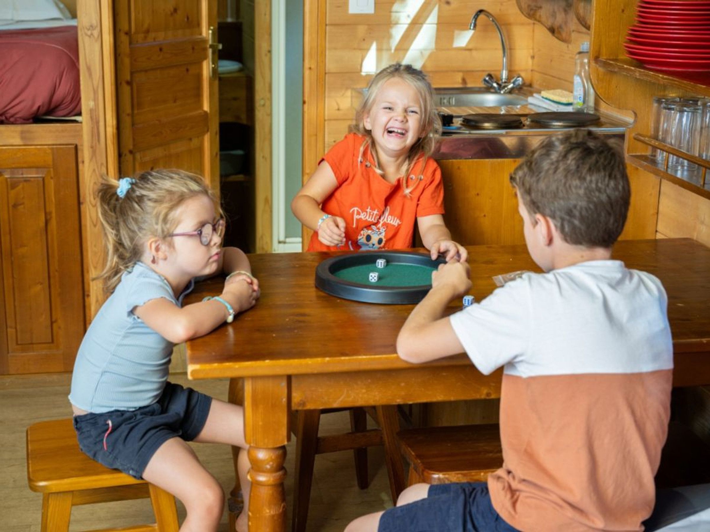 Three children playing a board game at a wooden table in a room with wooden walls and a kitchen area visible in the background.