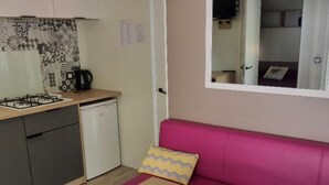 A compact kitchen with a gas stove, a small refrigerator, and a microwave. There's a pink sofa with a patterned cushion and a tray with bowls on a table in front of it. - Spacious mobile home with terrace in Nibelle (Nibelle)