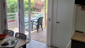 A room with a wooden table, chairs, a TV, and a sliding glass door leading to an outdoor area with a table and chairs. - Spacious mobile home with terrace in Nibelle (Nibelle)