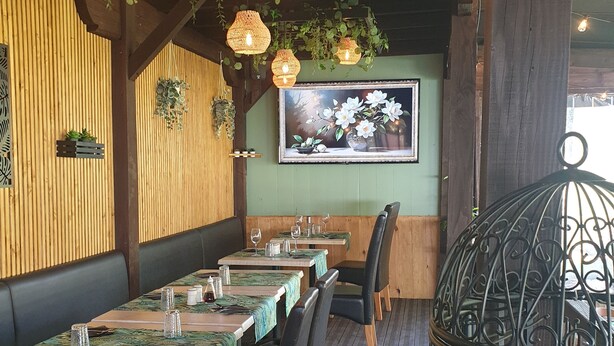 L’intérieur d’un restaurant présente des murs lambrissés en bois, des murs peints en vert et un grand tableau floral encadré. Les tables sont dressées avec des verres et des couverts, et des lampes en osier sont suspendues au plafond.