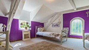 Une chambre avec un plafond en pente, des murs violets, un lit avec une literie à motifs floraux, une petite table de chevet et une fenêtre donnant sur un paysage verdoyant.