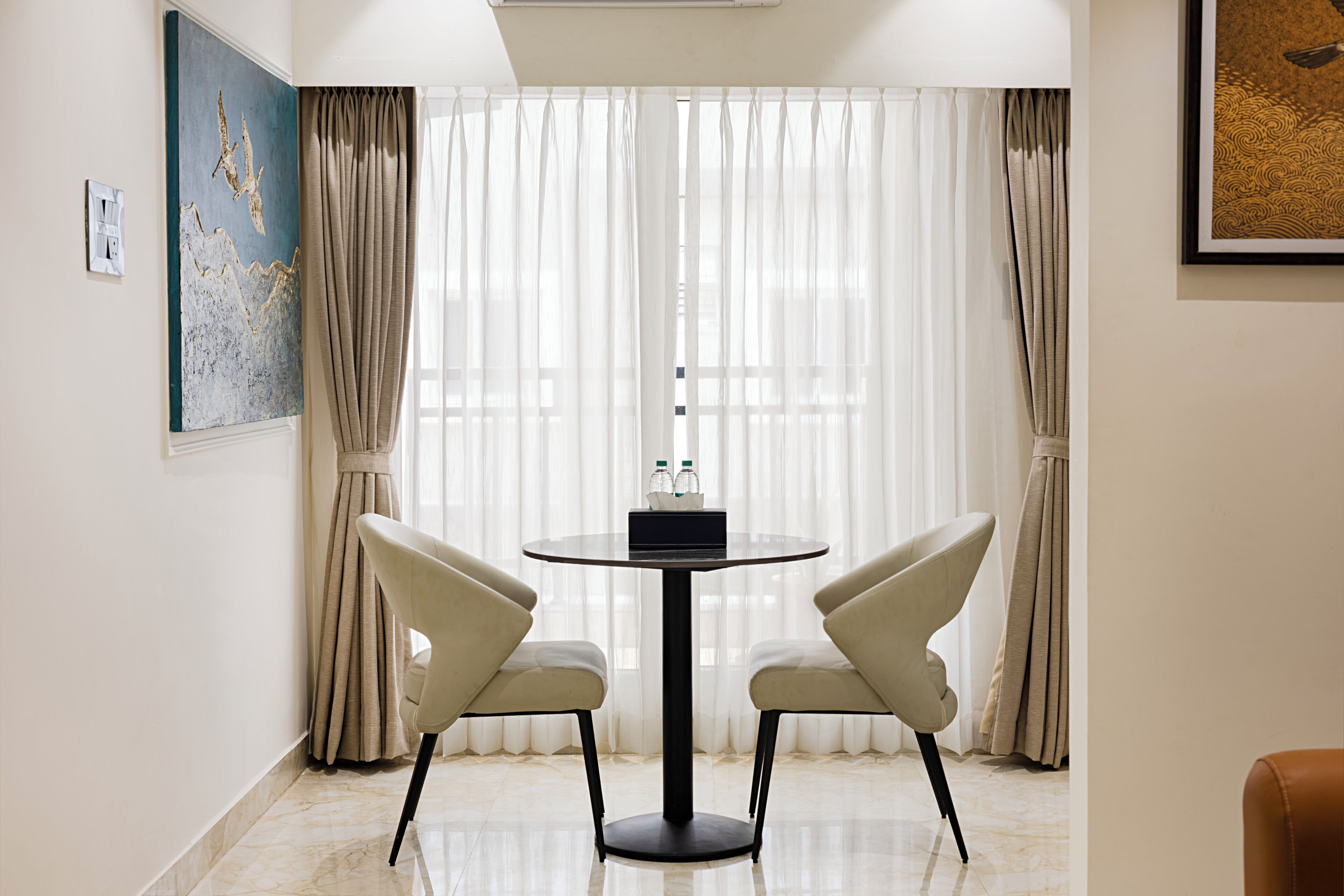 A room with a round table, two chairs, a painting on the wall, and a window with curtains.