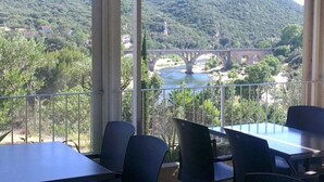 Zona d'estar a l'aire lliure amb taules i cadires, amb vistes a un riu i un pont, envoltada de vegetació.